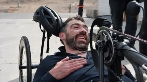 A man lies in a handcycle, with a helmet resting behind his head and team members nearby.