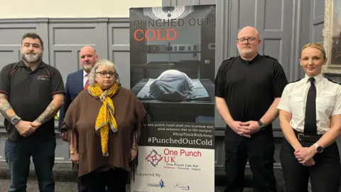 Andy Watson/BBC Three men and a woman, all representatives of Durham, Northumbria and Cleveland Police, are standing with Maxine Thompson-Curl in front of a sign which reads "Punched Out Cold" and has a picture of a body under a blue blanket on a mortuary slab. The man on the far left is wearing black, has dark hair and a greying beard. The man to his right is bald, has a beard and is wearing a blue suit with a tie. The man to his right is wearing black, is bald with a beard and has black glasses on. The woman, standing on the right, is wearing a white shirt and black tie which looks like a police uniform. She has blonde hair tied back. 