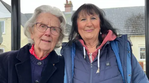 Two woman stood next to each other. They are both smiling and looking at the camera.