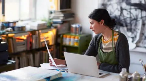 A young female artist sitting in her studio running her business from the laptop.