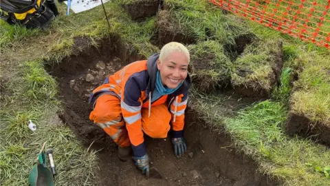 Grace McGrory/BBC Smiling archaeologist digging trench