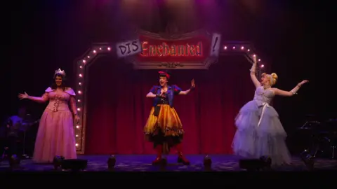 Three women on a stage. There is a red curtain backdrop and a sign that reads "Disenchanted!". The woman on the left has dark brown hair, a gold crown and a pink ballgown, dressed like Sleeping Beauty. In the middle is a woman with black hair, a red headband and a blue and red top with a gold skirt and red boots - dressed as Snow White. On the right is a woman with blonde hair in a bun and a long pale blue ballgown with matching gloves, dressed as Cinderella