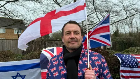 Nick smiles at the camera. He wears a jacket covered in union jack flags, he is holding an England flag and has two union flags behind him, as well as an American flag and an Israeli one.