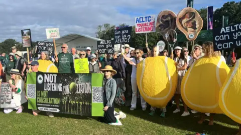 A large group of people, many holding banners and some dressed as tennis balls, hold up signs and banners, some saying 'You're not listening'.