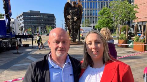 The parents of Connor Brown standing in front of the Knife Angel in Keel Square. His dad has a shaved head and goatee-style beard. His mum has blonde hair and wears a red top. 