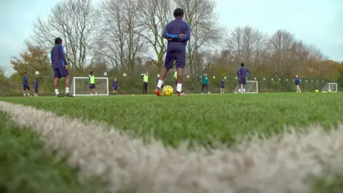 A football pitch, viewed from ground level. Several footballers, wearing matching shirts and shorts, are in the middle of a training session.