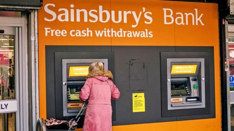 A woman using an ATM under a large Sainsbury's Bank sign