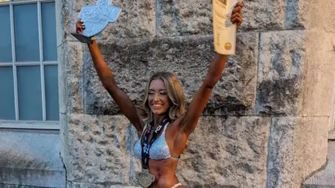 Mia Collins Mia Collins wearing a silver bikini and with a dark spray tan. She is standing with her arms in the air holding awards and wearing a medal round her neck.