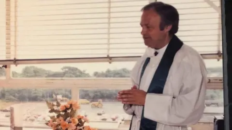 D J Photography A younger Anthony Pierce stands clasping his hands with his back to a window. He is side of frame and is wearing white robes with a dark silk stole around his neck. 