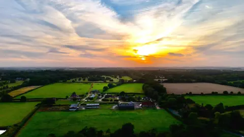 Shaun A setting sun shines through wispy clouds as the sky behind them graduates from orange to blue. The lower half of the picture is a green landscape of fields and trees with a farm and stables