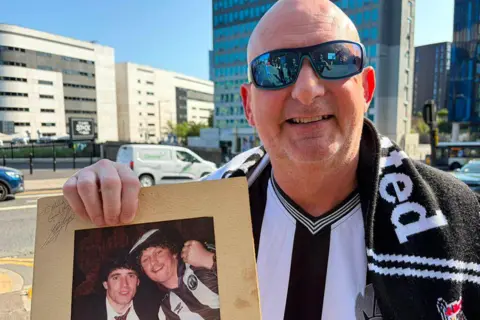 BBC/Adam Samuel Malcolm smiling into the camera while holding up an old framed photo of himself and Kevin Keegan where Malcolm is wearing a curly dark wig to match Keegan's hair. Malcolm is bald and wearing blue sporty sunglasses and an old Newcastle shirt and scarf. He is standing in front of St James' Park and the Strawberry Place road is behind him.