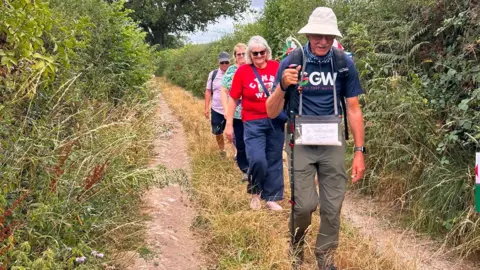 Andy Hockey John leads the way down a narrow country lane, with tall hedges either side. He holds a walking stick, with his family members walking in a line behind him. 