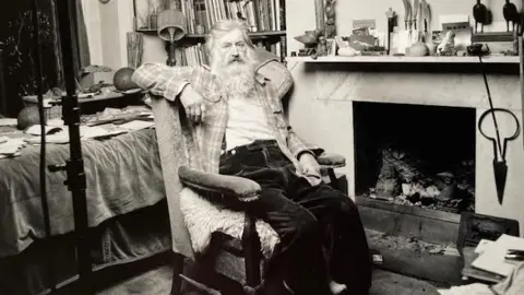 Norman McBeath RSA The sculptor Michael Black is pictured reclining in an old armchair in his home. The photo is black and white, and he sits in front of a large stone fireplace. He is wearing baggy black trousers, with a checked shirt over a white top. A bookshelf can be seen along with a surface littered with notebooks and other items