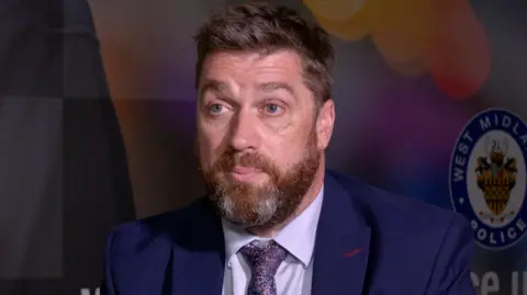 Mr George is wearing a navy suit and lilac shirt and tie. He is sat in front of police insignia on a background wall. He has a brown beard with white hairs in it and short brown hair