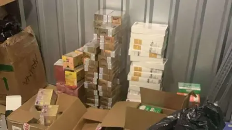 Suffolk Trading Standards Brown and white boxes of illegal vapes stacked on top of each other within a storage unit. Some smaller boxes sit in larger brown boxes. A black bin back can be seen to the right of the image. 