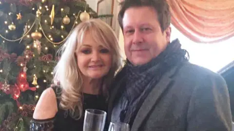 A glamourous blonde woman wearing a sequined black top, stood with her arm around a brunette man wearing a smartgrey coat and a scarf. They are both holding champaign glasses. Behind them is a Christmas tree decorated with baubles and lights.