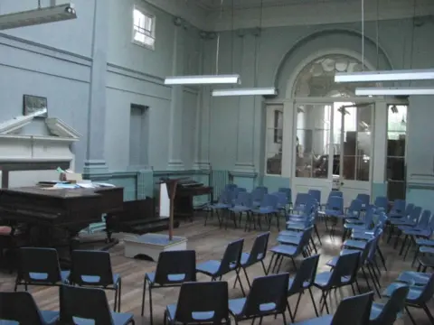 Andrew Leonard A room has a grand piano and lots of blue chairs arranged in a semi circle. It has blue painted walls and low-hanging strip lights. 