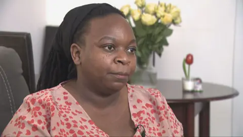 A woman with long black hair, tied back under a hair band, sitting on a sofa. She is wearing a pink top with a red pattern. There are flowers on a wooden table behind her. 