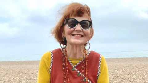 Marcia Riddington with red hair sits on a sea wall smiling at the camera. She wearing a yellow top with a red vest over the top and a skirt made up of a former quilt with different patches of fabric.