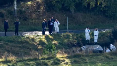 Police officers and forensics experts, who are wearing white overalls, at the site of the crashed car.