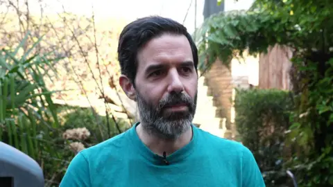 A man stands in a garden wearing a blue T‑shirt with a small microphone clipped to it. Behind him are bushes and trees.