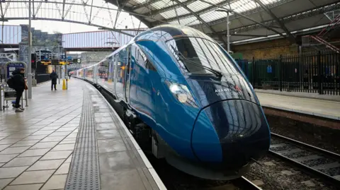 PA Media The front of a blue Avanti West Coast train next to a platform at Liverpool Lime Street station. A few people wait on the platform with a glass station roof arching over.