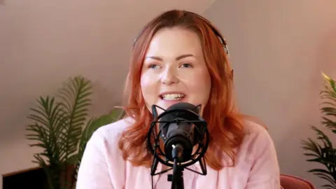 A woman, Lucy Edwards, has red hair and wears a pink jumper. She's sitting behind a microphone