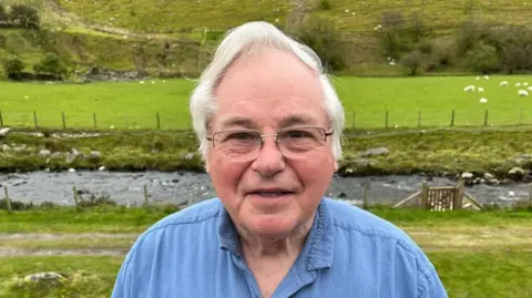 Robin Morris dengan kemeja biru dekat rumahnya di tepi sungai Ystwyth di Mid Wales. 