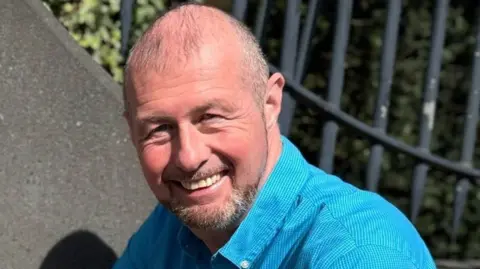 Justin Bellhouse is wearing a light blue, buttoned up shirt. He has thinning hair and a goatee beard. He is smiling.