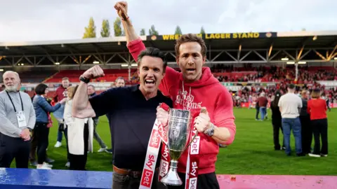 PA Media Hollywood stars Ryan Reynolds and Rob McElhenney celebrate with a trophy at the Wrexham AFC grounds, with people on the pitch behind them.
