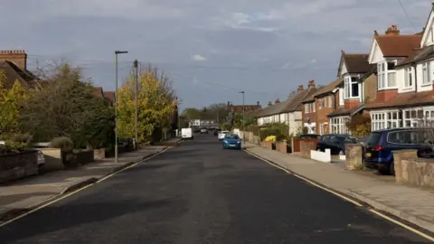 Facundo Arrizabalaga The road is newly resurfaced, black and wide. It has single yellow lines along the side. A few cars are parked on the road in the distance. 