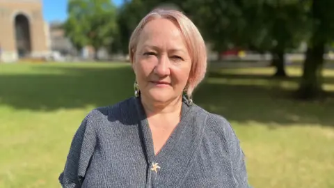 MP Kerry McCarthy wearing a grey wrap top, standing on College Green, a grassy area