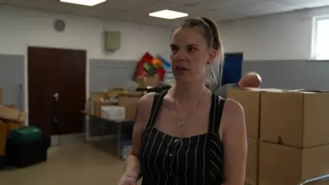 A woman with brown hair tied back and a black headband is wearing a black and white striped jumpsuit. There are brown boxes in a room behind her