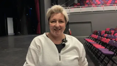 Pollyanne Trapmore-Shaw - a woman with short blond hair - stands in a large theatre auditorium with rows of red and purple seats to the right and a stage curtain to the left. She is wearing a white cardigan over a black top.