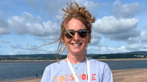A woman with a pale blue t-shirt displaying the logo of Sideshore, wearing sunglasses, hair tied up and stood in front of the beach and sea.