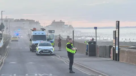 Phillip Norton/BBC Emergency vehicles along the seafront and an officer standing in the foreground. There are a number of buildings in the background and blue flashing lights can be seen.
