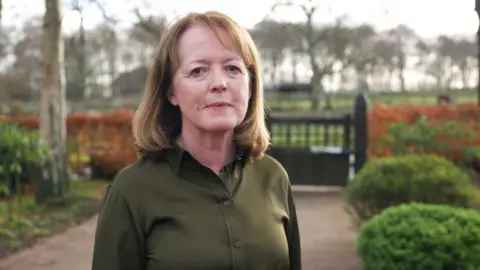 Dr Susan Gilby wears a dark green shirt and is standing on a path in front of a gate with red flowers and tall trees in the background