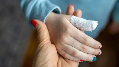 A close-up of an adult's hand with red painted nails, holding a child's hand with a bandage on the thumb