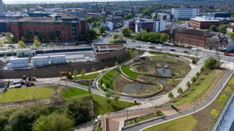 Riverside Park seen from the air. It has two ponds, landscaped grassland, young trees and walking paths. It is on the edge of Sunderland city centre, with Sunderland University's campus visible in the distance, as well as a Premier Inn. 