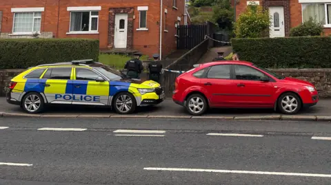 A photo taken from the opposite side of a road showing a parked police car outside a row of semi-detached, red brick houses in north Belfast. Three uniformed officers are standing with their backs to the camera. A red car is also parked blocking the driveway of the house. A cordon is placed along the bottom and mid-way up the driveway of the house.