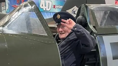 An elderly man is sitting in the cockpit of a Spitfire, wearing a peaked cap and saluting while he looks at the camera