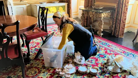 BBC/Rachel Royce A woman is unpacking delicate china while kneeling on the floor of an ornate, traditionally decorated room with patterned red-and-gold carpet and antique furniture. She is reaching into a large clear plastic storage box surrounded by wrapped items and neatly arranged teacups, saucers and plates. There are gold-framed furnishings, draped curtains and decorative side tables in the background.