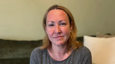 A woman wearing grey sits on her green couch with pillows behind her 