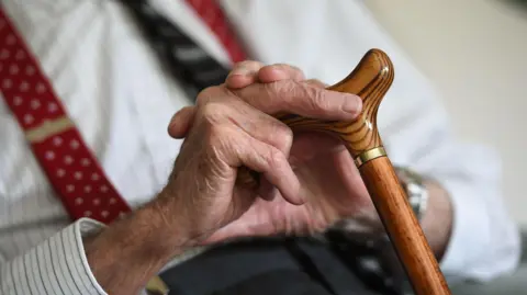 An elderly man holding a wooden walking stick. The man is wearing a striped shirt and a red tie. His hands are wrinkled.