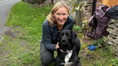 Handout Tara knelt down on a path with her dog smiling at the camera. Her walking backpack is to the side next to a bowl of water for her dog.