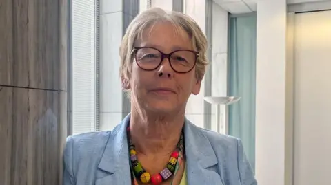 Jane Ashworth wears a light blue jacket, with a bold colourful necklace. She is wearing glasses and smiling at the camera. 