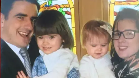 Handout A family photograph showing a man with black hair and wearing a tie and shirt holding his child, while his wife, who is wearing thick-rimmed glasses, holds their older child.