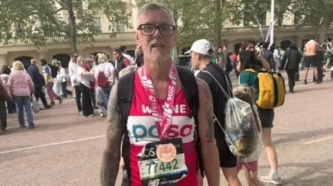 Wayne has grey hair and black glasses and is wearing a pink vest with white writing that reads "Wayne, PDSA". His race bib is black with green numbers on it. He has a medal around his neck which has a pink and white ribbon with a bronze coloured medal