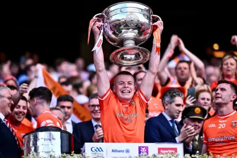 Getty Images Aidan Nugent, a dark haired man in an orange Armagh GAA top, lifting the large, silver Sam Maguire trophy above his head with both hands
There is a large crowd of fans and teammates cheering in the background.
The former taoiseach Simon Harris, a grey haired man in a navy suit, is pictured beside him.
