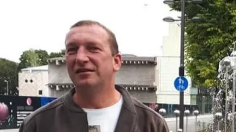 A man with short brown hair which is receding slightly. He is wearing a brown jacket over a white t-shirt